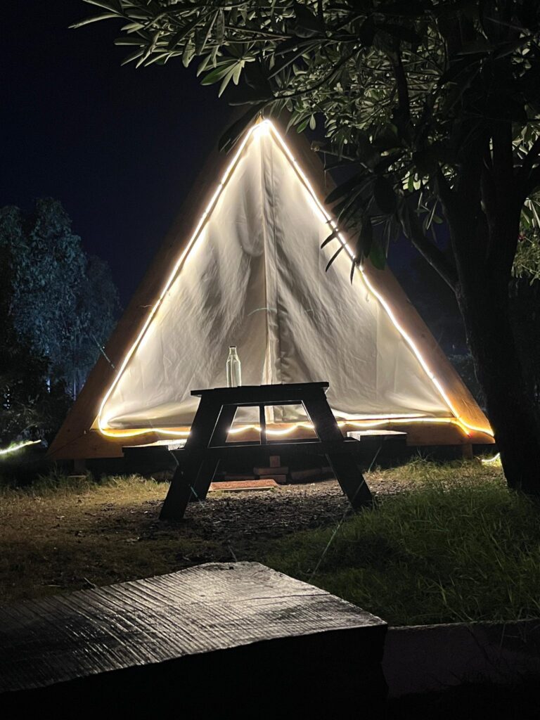 A cozy triangular tent illuminated by soft lights at night, set in a grassy area surrounded by trees.